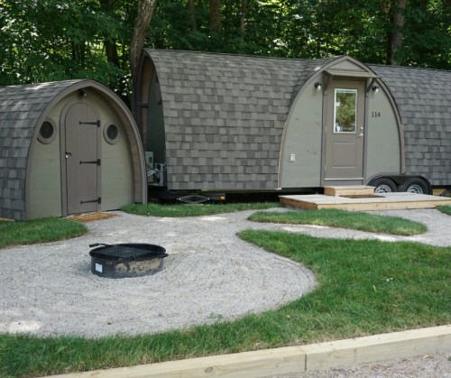 Two unique, curved cabins with a gravel path and fire pit in a wooded area. Green grass surrounds the cabins.