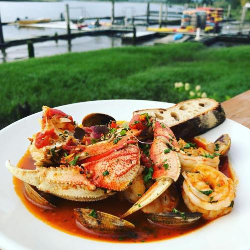A bowl of seafood stew with crab, shrimp, and clams, garnished with herbs, served with a slice of bread.