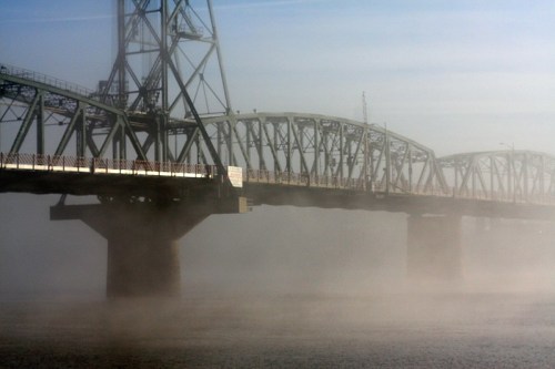 A foggy view of a steel bridge spanning a river, with mist rising from the water below.