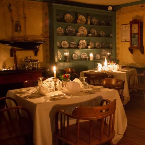 Cozy restaurant interior with candlelit tables, vintage decor, and shelves displaying decorative plates.