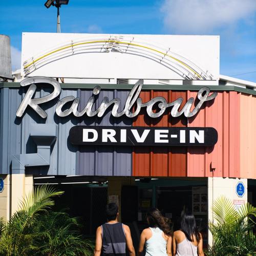 A colorful sign for "Rainbow Drive-In" with three people walking towards the entrance under a blue sky.
