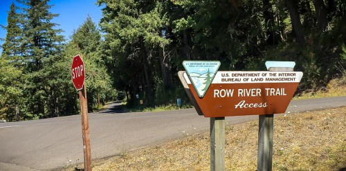Sign for Row River Trail Access next to a stop sign, surrounded by trees along a paved road.