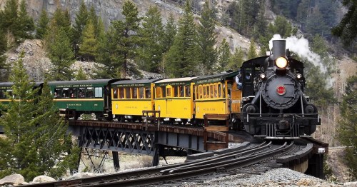 A vintage steam train with colorful carriages travels along a curved track through a forested landscape.