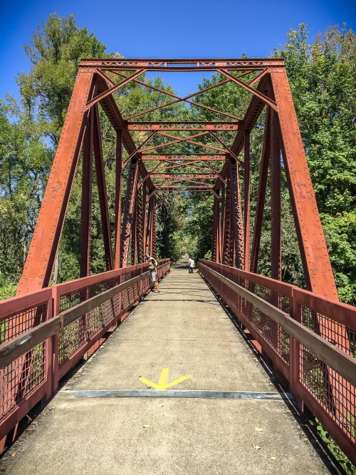A red metal bridge spans a path surrounded by green trees under a clear blue sky.