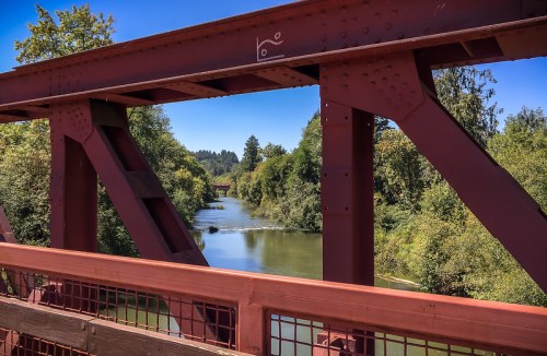 A view of a river framed by a red metal bridge, surrounded by lush greenery under a clear blue sky.