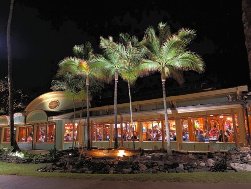 A brightly lit restaurant at night, surrounded by palm trees and a small pond, with diners visible through large windows.