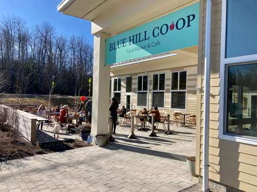 Exterior of Blue Hill Co-op with outdoor seating, people dining, and a sign above the entrance on a sunny day.