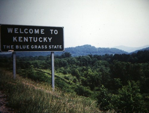 A welcome sign for Kentucky, "The Blue Grass State," with green hills and trees in the background.