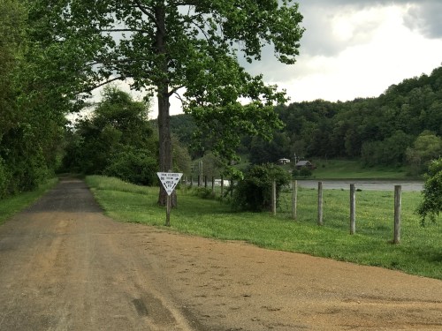 A gravel path lined with trees leads to a river, with a warning sign visible along the way.
