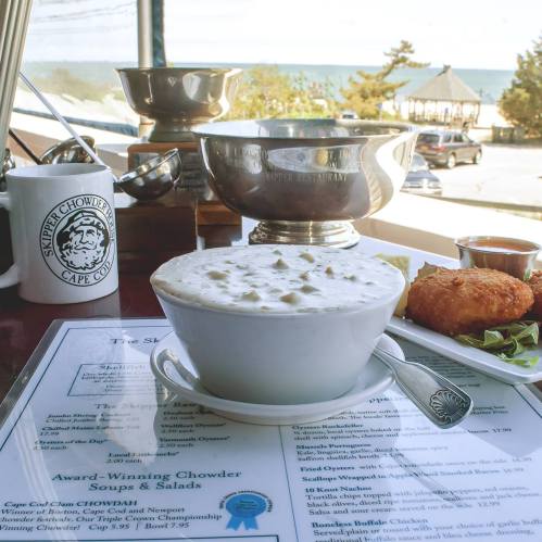 A bowl of clam chowder on a table with a menu, a crab cake, and a view of the beach in the background.