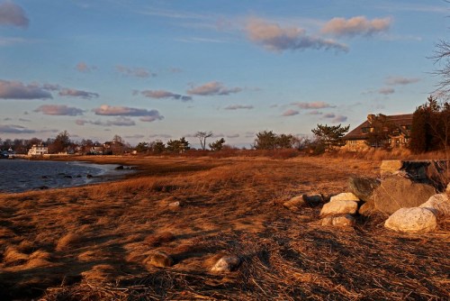 A serene coastal scene at sunset, featuring a grassy shore, scattered rocks, and a distant house under a cloudy sky.