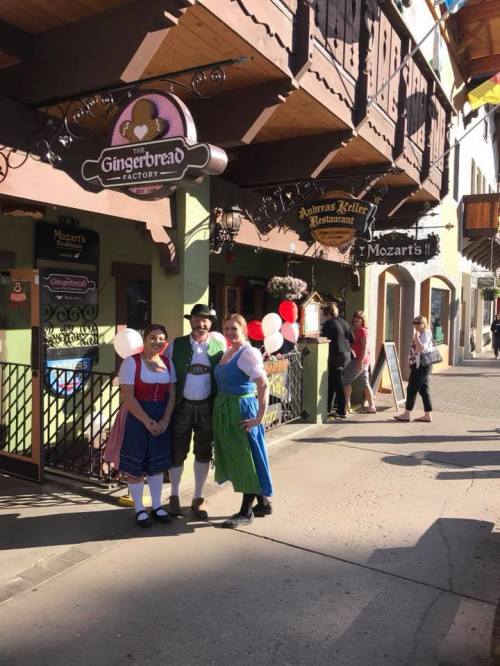 Three people in traditional costumes stand outside a charming storefront with festive decorations and balloons.