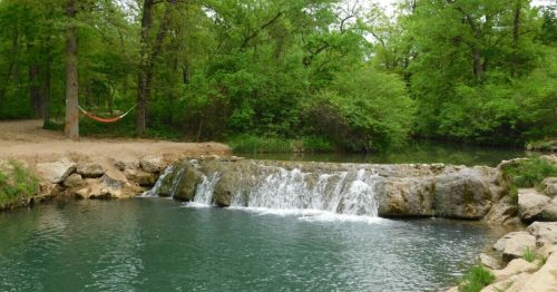 A serene natural scene featuring a small waterfall flowing into a calm pool, surrounded by lush green trees.