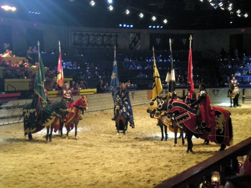 Knights in colorful armor on horseback, holding flags, in a medieval arena with an audience in the background.