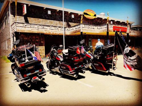 Three motorcycles parked in front of a rustic building, adorned with flags and gear, under a clear blue sky.