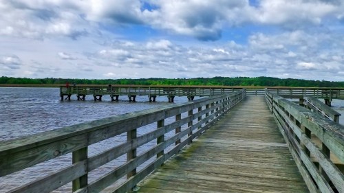 A wooden pier extends over calm water, surrounded by lush greenery and a cloudy blue sky.