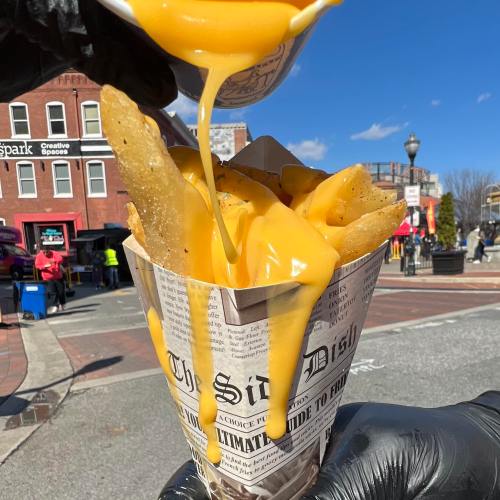 A hand pours melted cheese over a cone of crispy fries served in a newspaper-style container.
