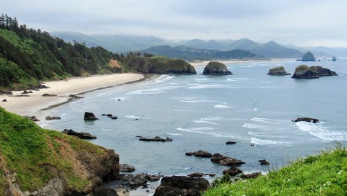 A scenic coastal view featuring a sandy beach, rocky outcrops, and rolling waves under a cloudy sky.