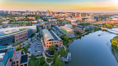 Aerial view of a city skyline at sunset, featuring buildings, a river, and green spaces along the waterfront.