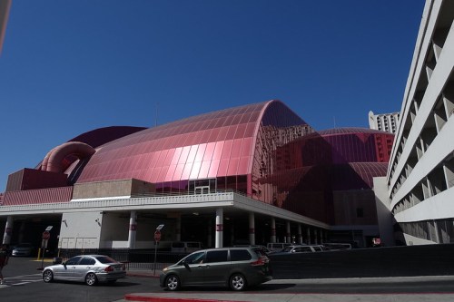 A large, modern building with a shiny pink exterior under a clear blue sky, surrounded by parked cars.