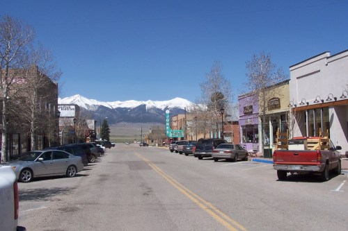 A small town street lined with shops and parked cars, with snow-capped mountains in the background under a clear blue sky.