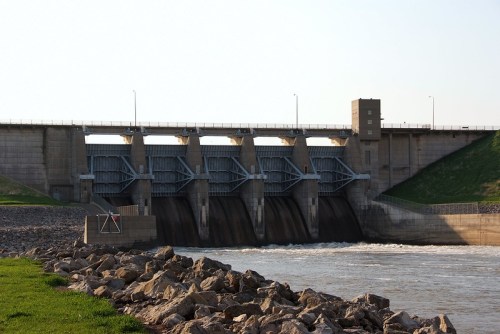 A concrete dam with multiple gates, water flowing beneath, and grassy banks on either side.