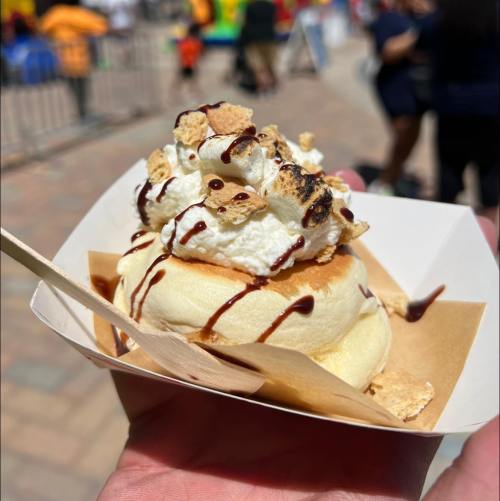 A hand holding a dessert with whipped cream, chocolate drizzle, and crushed graham crackers in a paper cup.