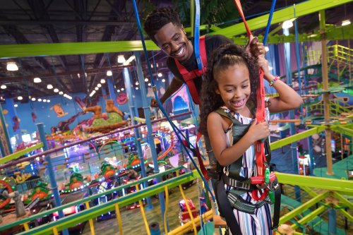 A man and a girl enjoy a fun indoor amusement park, harnessed in safety gear while navigating colorful ropes.