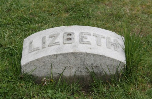 A weathered gravestone with the name "Lizbeth" engraved on it, surrounded by grass.