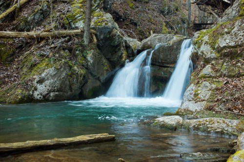 A serene waterfall cascades over rocks into a clear pool, surrounded by lush greenery and moss-covered stones.