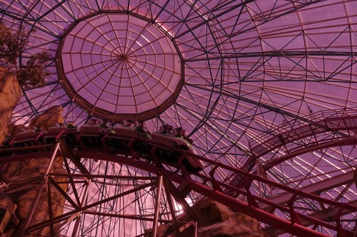 A roller coaster twists through a vibrant, glass-roofed structure with a purple hue.