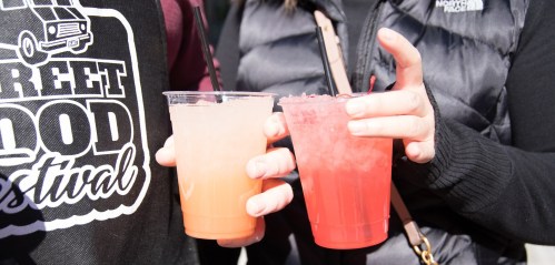 Two hands hold colorful drinks with straws at a street food festival, one orange and one pink.