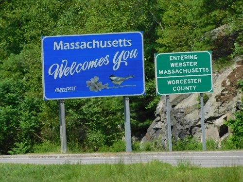 A welcome sign for Massachusetts with a bird and flowers, alongside a sign indicating entry into Webster, Worcester County.