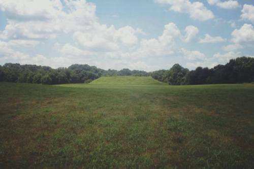 A grassy field with a large, flat-topped mound in the distance, surrounded by trees and a blue sky with clouds.