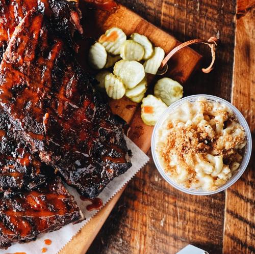 A wooden platter with BBQ ribs, pickles, and a cup of macaroni and cheese topped with breadcrumbs.