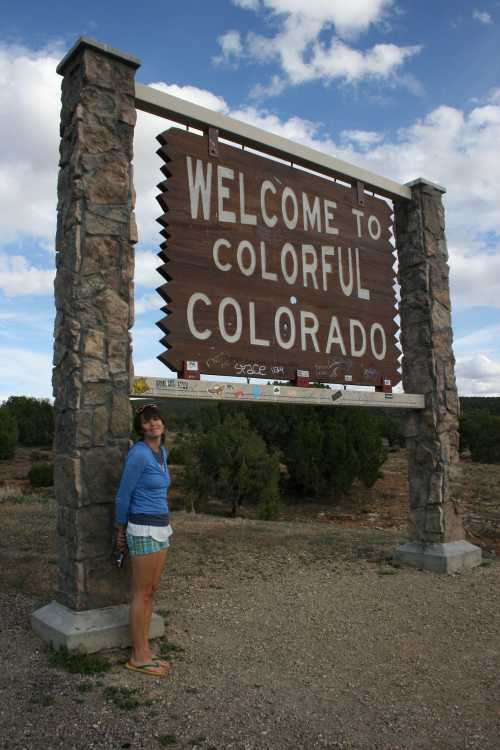 A person stands beside a large "Welcome to Colorful Colorado" sign in a rural landscape with trees and clouds.