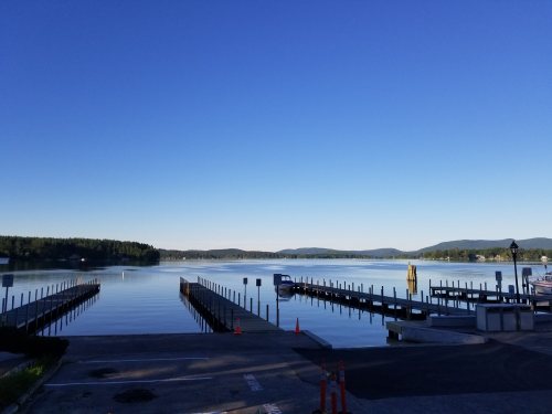 A calm lake scene with docks extending into the water, surrounded by trees and mountains under a clear blue sky.