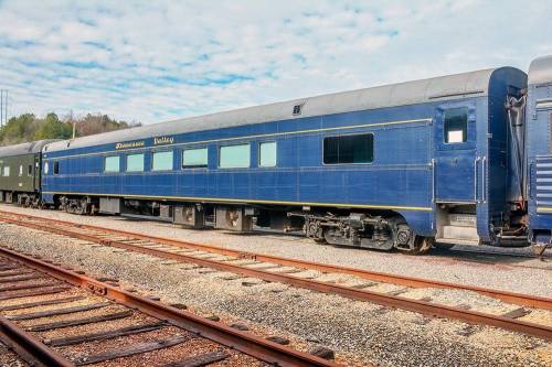 A blue vintage train car parked on a railway track, surrounded by greenery and a cloudy sky.