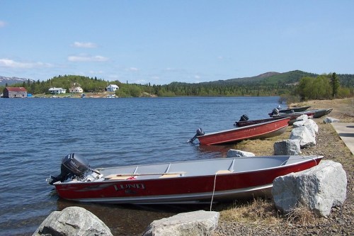 Four red boats are moored by a rocky shore, with a calm lake and green hills in the background under a clear blue sky.