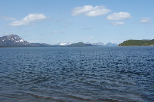 A serene view of a calm lake surrounded by mountains and a clear blue sky with scattered clouds.