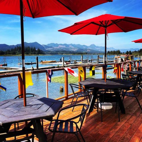 A scenic lakeside view with red umbrellas over wooden tables, surrounded by mountains and colorful flags.