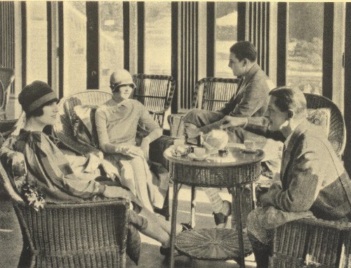 A vintage scene of four people in stylish attire seated in a sunlit room with wicker furniture and drinks on the table.