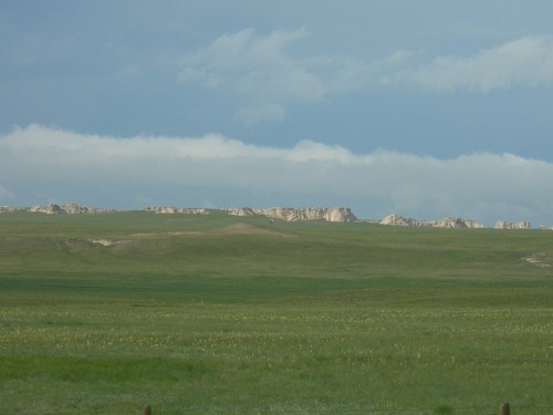 A vast green landscape with rolling hills and rocky outcrops under a cloudy blue sky.