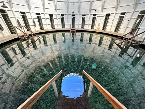 A circular indoor pool with clear water, surrounded by wooden steps and white walls, reflecting light and shadows.