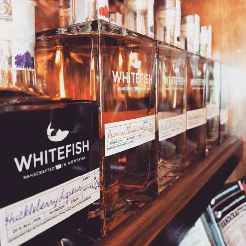 A shelf displaying various bottles of Whitefish handcrafted spirits, labeled with flavors and batch information.
