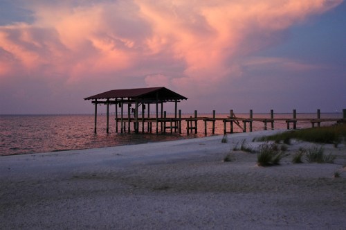 A serene beach scene at sunset, featuring a wooden pier extending into calm waters under colorful clouds.