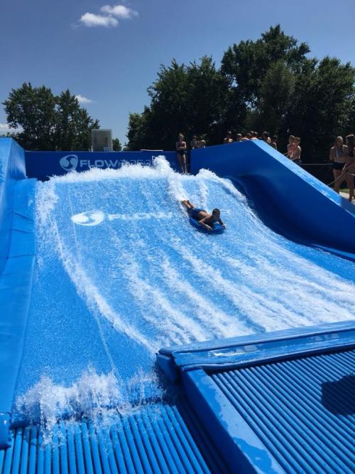 A person slides down a blue water ramp, surrounded by spectators and trees on a sunny day.