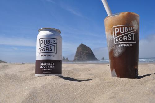 A can of root beer and a glass of root beer float on a sandy beach with a rocky formation in the background.