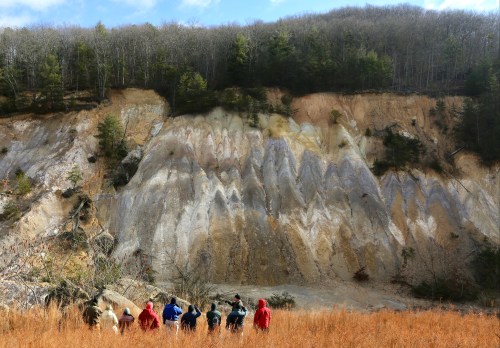 A group of people in colorful jackets stands in a field, facing a layered, rocky hillside with trees above.