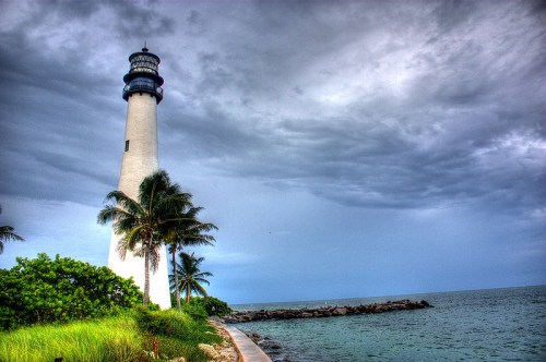 A tall lighthouse stands by the shore, surrounded by palm trees and a cloudy sky over the ocean.